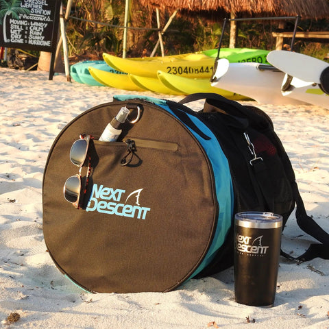 Next Descent bag with tumbler and sunglasses on sandy beach near kayaks