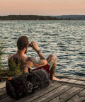 Man sitting on dock with 90L Large Mesh Duffle Bag For Scuba Gear Snorkel Equipment And Water Sports Adventure beside him.