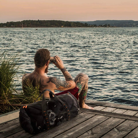 Man sitting on dock with 90L Large Mesh Duffle Bag For Scuba Gear Snorkel Equipment And Water Sports Adventure beside him.