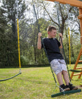 Child standing on green stand up swing for backyard playsets with 14x14 inch base, coated chains, attached to wooden playset