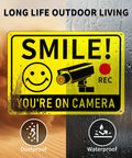 Smile You're On Camera Signs Two Pack Outdoor Video Surveillance Warning Signs 10x7 Rust Free Aluminum UV Printed Weatherproof Security Sign in desert and rain setting