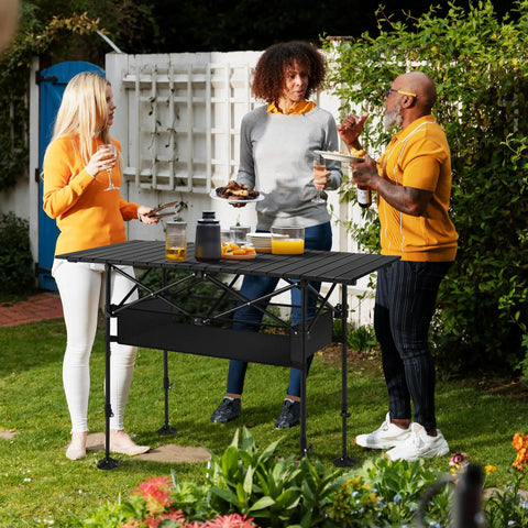 Three people around Portable Adjustable Aluminum Camping Table With Built In Storage For Grill BBQ Travel Picnic Beach And Backyard Use Lightweight Outdoor Table Supports Up To 110 Lbs And Folds Flat For Easy Carry in a garden setting.
