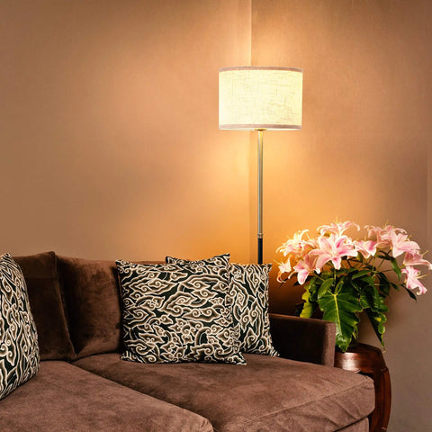 Emery LED Floor Lamp With Drum Shade illuminating a cozy living room corner next to a brown sofa with patterned cushions and flowers.