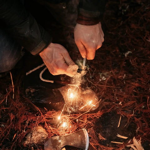 Hands using Ferro Rod Fire Starter Survival Tool With Wood Handle, Up To 15,000 Strikes, Three Eighths Inch Fire Steel, Multi Tool Striker And Lanyard, creating sparks on forest ground.