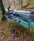 Person relaxing in Forest Green Hybrid Ground Tent Or Hammock With Strap Bundle For Car Camping And Backpacking by a forest stream