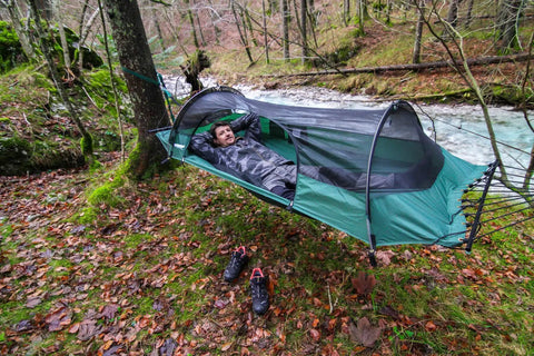 Person relaxing in Forest Green Hybrid Ground Tent Or Hammock With Strap Bundle For Car Camping And Backpacking by a forest stream