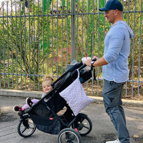 Man pushing stroller with child and Waterproof Wet Dry Bag Set for Cloth Diapers and Swimwear attached, urban park setting.