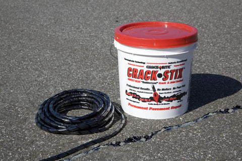 Coiled crack filler with red-lidded bucket on asphalt, labeled 125 Feet Of Blacktop Crack Filler For Asphalt And Concrete Weather Resistant No VOCs