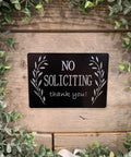 Black "No Soliciting" sign on a wooden background surrounded by green leaves.
