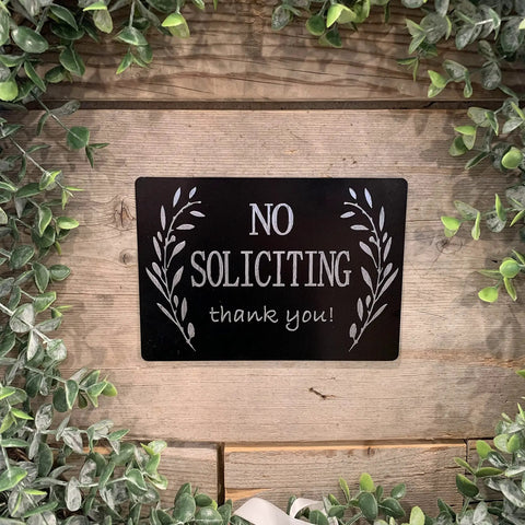 Black "No Soliciting" sign on a wooden background surrounded by green leaves.
