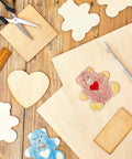 Basswood craft sheets 12x18x1/8in with assorted wooden cutouts (heart, bear, cloud) and tools on a worktable.
