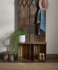 Rustic wooden coat rack with hooks, hanging hat and jacket. Plant and shoes on bench below, with a wire basket on the side.