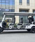 White Eight Passenger Electric Golf Cart Rolled Up Tight With Beefy Dual Arm Suspensions Silky Smooth Five Kilowatt Motor Plus Touchscreen Audio And Deep Reclining Lounge Seats parked on pavement with modern building in background.