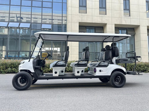 White Eight Passenger Electric Golf Cart Rolled Up Tight With Beefy Dual Arm Suspensions Silky Smooth Five Kilowatt Motor Plus Touchscreen Audio And Deep Reclining Lounge Seats parked on pavement with modern building in background.