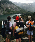Three hikers with backpacks overlooking a mountain lake, camping gear visible, showcasing outdoor adventure.