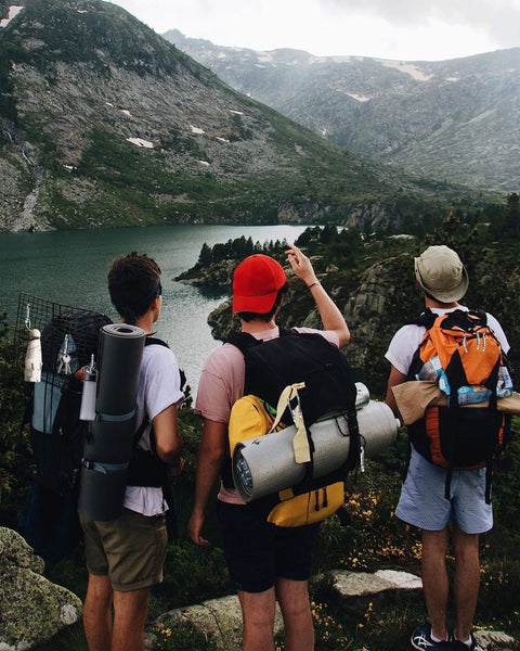 Three hikers with backpacks overlooking a mountain lake, camping gear visible, showcasing outdoor adventure.