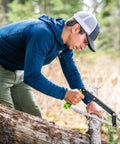 Person using Folding Camp Saw With 12 Inch Blade For Backpacking, Hiking, Camping And Bushcraft, cutting wood with bright green accents.