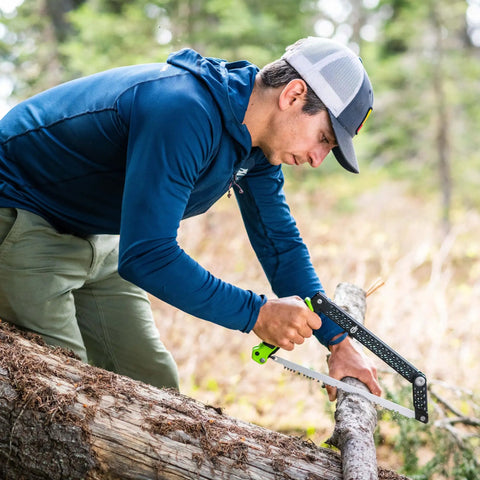 Person using Folding Camp Saw With 12 Inch Blade For Backpacking, Hiking, Camping And Bushcraft, cutting wood with bright green accents.