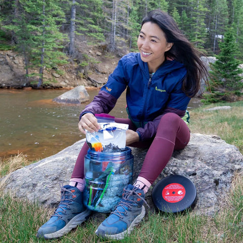 Person using BV475 Trek Portable Bear Resistant Canister For Backpackers next to a river while camping outdoors