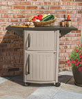 Outdoor Grilling Prep Station With Storage Portable Backyard Kitchen Counter Durable Resin Weather Resistant Top Cabinet For Serving Tray And More With Drop Leaf Extensions Taupe Brown in a garden setting with food items on top.