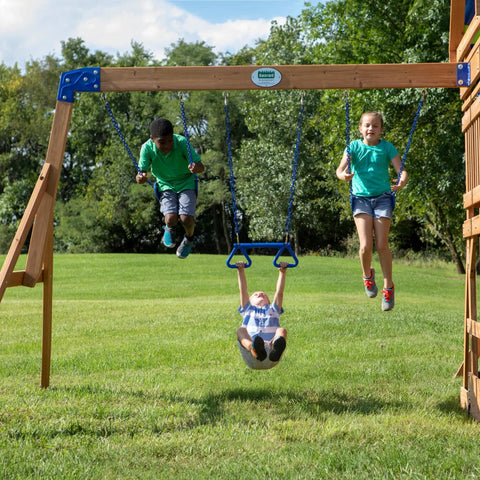 Children playing on the Beachfront Cedar Wood Swing Set With Wave Slide Elevated Deck Canopy Monkey Bars Rock Wall Ladder Shaded Picnic Area Snack Bar Window Belt Swings Trapeze Sandbox Playset For Kids in a backyard.