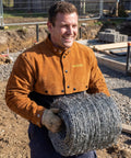 Rugged Full Grain Cowhide Welding Cape Sleeve With Kevlar Stitched Panels And Flame Resistant Barrier Against Flying Sparks And Hot Slag Shop Ready And Built To Go The Distance worn by a worker holding barbed wire on a construction site.