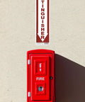 Go-To Durable Aluminum Fire Extinguisher Sign With Bold Red Fire Extinguisher Text And Downward Arrow 18 Inch Long By 4 Inch High Reflective White Background Office Hallway Safety Sign next to a red fire extinguisher box mounted on a wall.