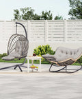 Cozy Outdoor Egg Swing Chair With Stand And Footrest Deep Cushioned Hammock Style Seat For Patio Or Indoor Use Waterproof Rattan Sturdy Metal Frame Comfy Lounge Chair In Dark Gray next to a matching lounge chair on a patio.