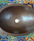 16 By 13 Inch Oval Skirted Vessel Copper Sink Hand Hammered Pure Copper Brown Finish For Modern Bath Semi Recessed Vessel One And A Half Inch Drain Opening Eighteen Gauge Handmade In Mexico CPS10 shown from top on colorful tiles.
