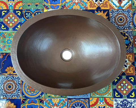 16 By 13 Inch Oval Skirted Vessel Copper Sink Hand Hammered Pure Copper Brown Finish For Modern Bath Semi Recessed Vessel One And A Half Inch Drain Opening Eighteen Gauge Handmade In Mexico CPS10 shown from top on colorful tiles.