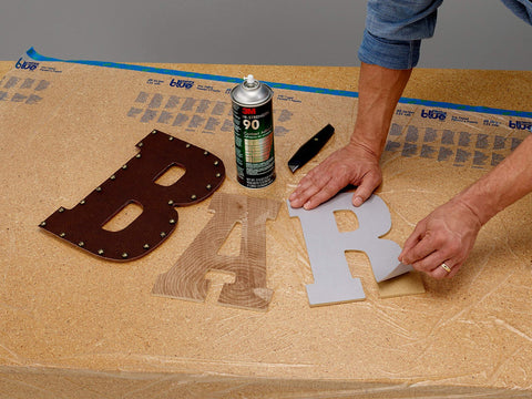 Person using Hi-Strength Spray Adhesive 90, Low VOC 14.6 oz can, CARB compliant, on wooden letters on a table.