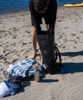 Person packing 25L Waterproof Surf Backpack For Adventure With Roll Top Closure Keeps Gear Dry In Any Weather on beach.