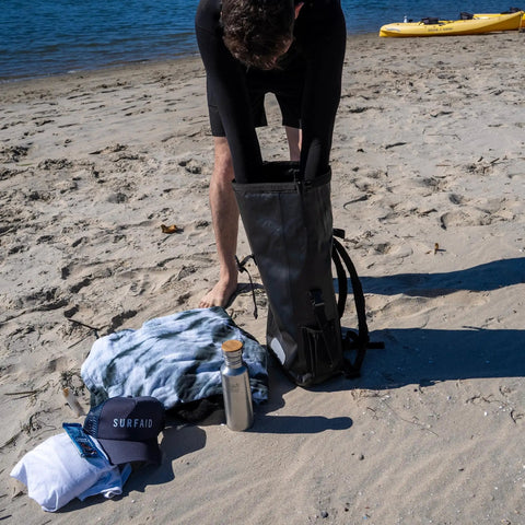 Person packing 25L Waterproof Surf Backpack For Adventure With Roll Top Closure Keeps Gear Dry In Any Weather on beach.