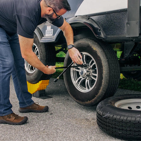 Man using Curved Trailer Aid With Wheel Chock And Rubber Base Pad, Six And A Half Inch Lift, Fits Trailers Up To Twenty Thousand Pounds And Tires Up To Thirty Two Inches, No Jack Needed For Easy RV And Camper Tire Changes Made In USA to change trailer tire