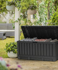92 Gallon Resin Deck Box In Dark Gray For Outdoor Storage Keep Patio Cushions And Throw Pillows Garden Tools Pool Floats Neat And Ready To Grab On The Patio Or In The Garage, open with pillows inside, surrounded by garden