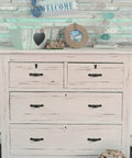 Wooden dresser with four drawers in a coastal-themed room, decorated with beach-inspired items and a welcome sign.