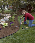 Person installing No Dig Landscape Edging Kit, 100 Ft Flexible Black Border With 32 Anchoring Spikes, around garden area.