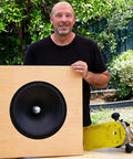 Man holding wooden panel with large speaker outdoors next to skateboard.