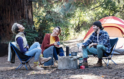 Three people sitting in a forest campsite with chairs and a tent, enjoying a laugh and holding mugs.