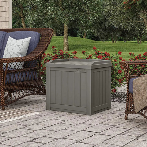 22 Gallon Outdoor Storage Box With Seat In Stone Gray Resin on patio, surrounded by wicker furniture in a garden setting
