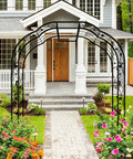 Tall Black Garden Arch Trellis 8.2 Ft High 6.2 Ft Wide Outdoor Arbor For Climbing Plants Roses And Vines Built To Last Perfect Lawn Wedding Archway Durable Weatherproof Construction in front of a house entrance with flowers lining the path under the arch.