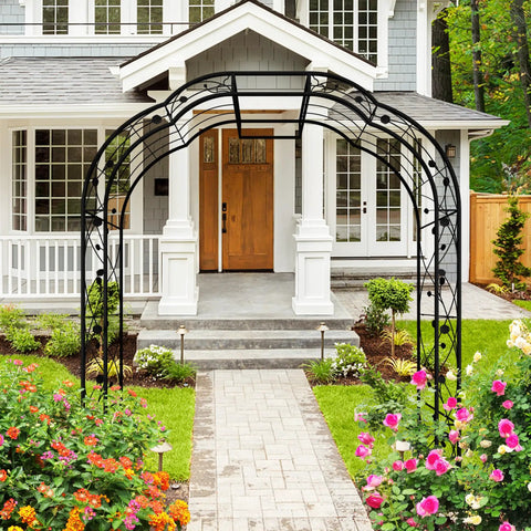 Tall Black Garden Arch Trellis 8.2 Ft High 6.2 Ft Wide Outdoor Arbor For Climbing Plants Roses And Vines Built To Last Perfect Lawn Wedding Archway Durable Weatherproof Construction in front of a house entrance with flowers lining the path under the arch.