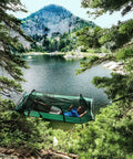 Person relaxing in a Forest Green Hybrid Ground Tent Or Hammock With Strap Bundle For Car Camping And Backpacking, near a mountain lake.