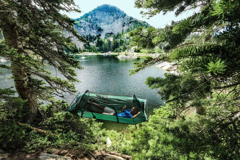 Person relaxing in a Forest Green Hybrid Ground Tent Or Hammock With Strap Bundle For Car Camping And Backpacking, near a mountain lake.