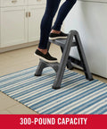 Two Step Folding Foot Stool 14 Inch High Gray Lightweight 300 Pound Capacity in use in a kitchen setting by an adult.
