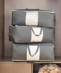 Three grey Roomy 105L Extra Large Storage Bags For Blankets Bedding And Clothes 3 Pack With 4 Handles stacked in closet with clear window.
