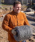 Man wearing Leather Welding Jacket With Open Back And Cape Sleeve, Bib Apron Style Welder Coat, holding barbed wire on construction site.
