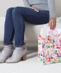 Person sitting next to 10.47 Inch Square Gift Bag With Watercolor Dots And Just For You Design, sturdy paper bag