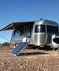 130W Portable RV Solar Panel Kit With Folding Panels set up outside a silver RV, showing adjustable legs and battery clamps for off-grid power