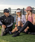 Three people sitting on grass wearing athletic wear and caps, smiling and conversing outdoors on a rooftop.